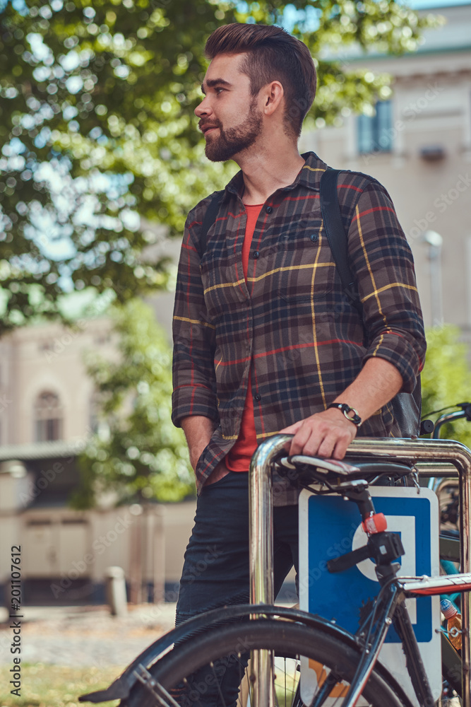A handsome fashionable young male with stylish haircut and beard, wearing fleece shirt, stands in the parking lot for bicycles in the park.