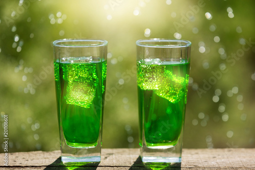 two tall glasses with ice and a green sparkling drink, against a background of greenery