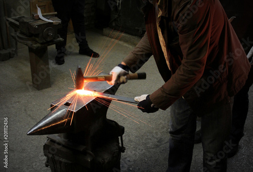 The blacksmith manually forging the molten metal on the anvil in smithy with spark fireworks. Spark. Dark background.