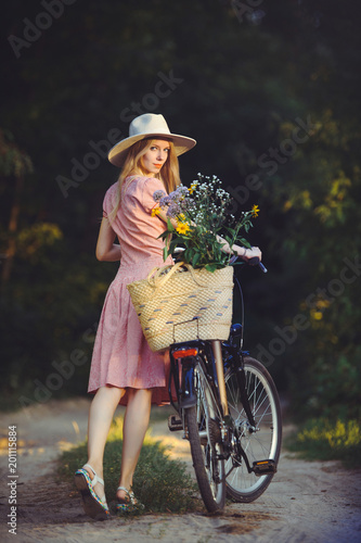 Beautiful girl wearing a nice pink dress having fun in a park with a bicycle holding a beautiful basket with flowers. Vintage landscapes. Pretty blonde with retro look, bicycle and basket with flowers