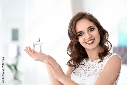 Beautiful young bride with bottle of perfume on blurred background