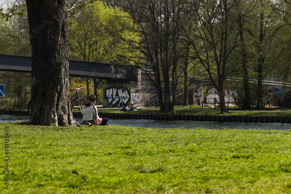 Fototapeta premium Parkwiese im Frühling, Frau sitzt auf einer Wiese am Wasser, frische grüne Parkwiese, Schöneweide, Kaisersteg, Frühjahr