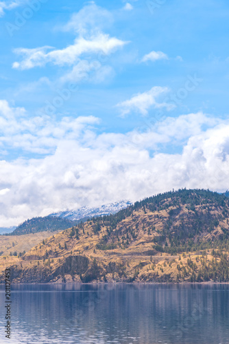 Landscape of lake, grassy hills, snow covered mountains, blue sky and clouds on sunny day © Amy Mitchell