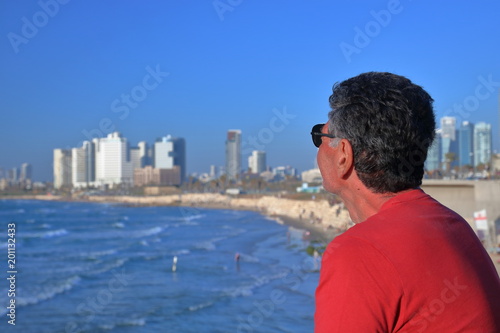Satrszy mężczyzna z lekko siwiejącymi włosami, w czerwonej koszulce i okularach słonecznych, stoi bokiem i spogląda na panoramę zatoki Morza Śódziemnego, plażę i wyokie, nowoczesne budynki Tel Awiwu