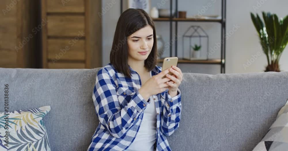 Beautiful young woman in the blue plaid shirt holding a smartphone and typing on its screen at home. Indoor Beautiful young woman in the blue plaid shirt holding a smartphone and typing on its screen at home. Indoor