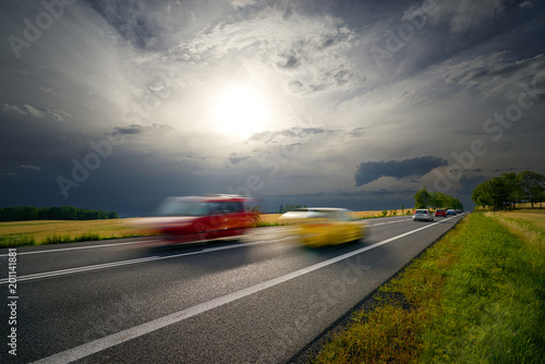 Motion blurred cars driving on a road in a rural landscape under the sun shining through the clouds before the storm