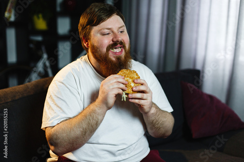Smiling fat man eats burger sitting before a TV-set