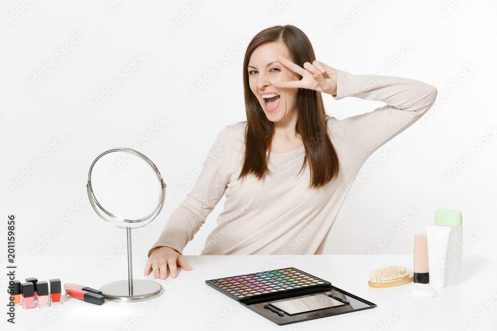 Happy fun young woman showing victory sign, sitting at table applying makeup with set facial decorative cosmetics isolated on white background. Beauty female fashion lifestyle concept. Area copy space Happy fun young woman showing victory sign, sitting at table applying makeup with set facial decorative cosmetics isolated on white background. Beauty female fashion lifestyle concept. Area copy space