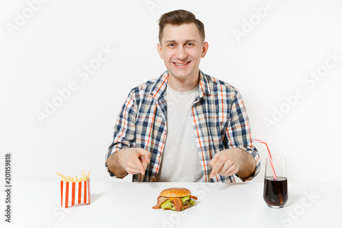 Handsome young man in shirt sitting at table with burger, french fries, cola in glass isolated on white background. Proper nutrition or American classic fast food. Advertising area with copy space.