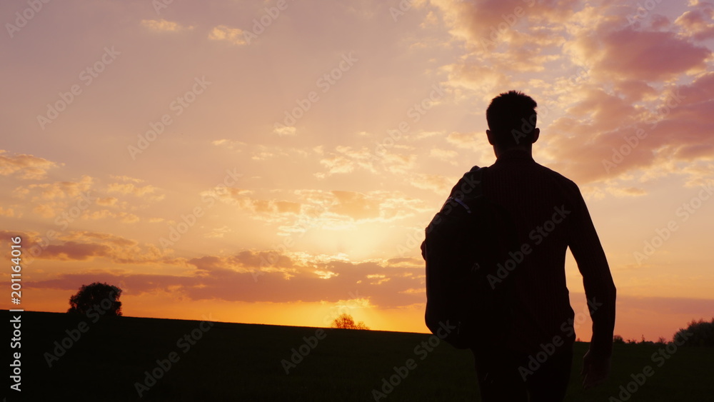 A teenager with a backpack over his shoulder goes towards the sunset in the field or in the countryside. Concept - new research, forward to the unknown, to leave home