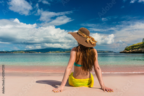 Rear view of a young woman with straw hat looking at an idyllic view while sitting on the sand at Pink Beach in Komodo Island, Indonesia