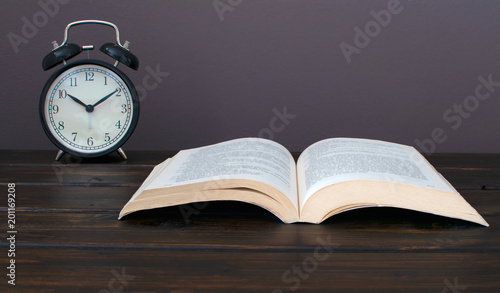 Still life photo of an open book with black alram clock on wooden table