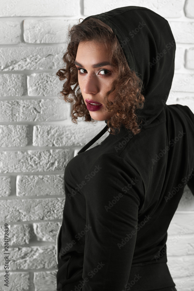 Backview of passionate curly model looking at camera. girl wearing black hood posing near white wall. having beatiful young face, intresting hairstyle with curles. Woman wearing black sportsuit.