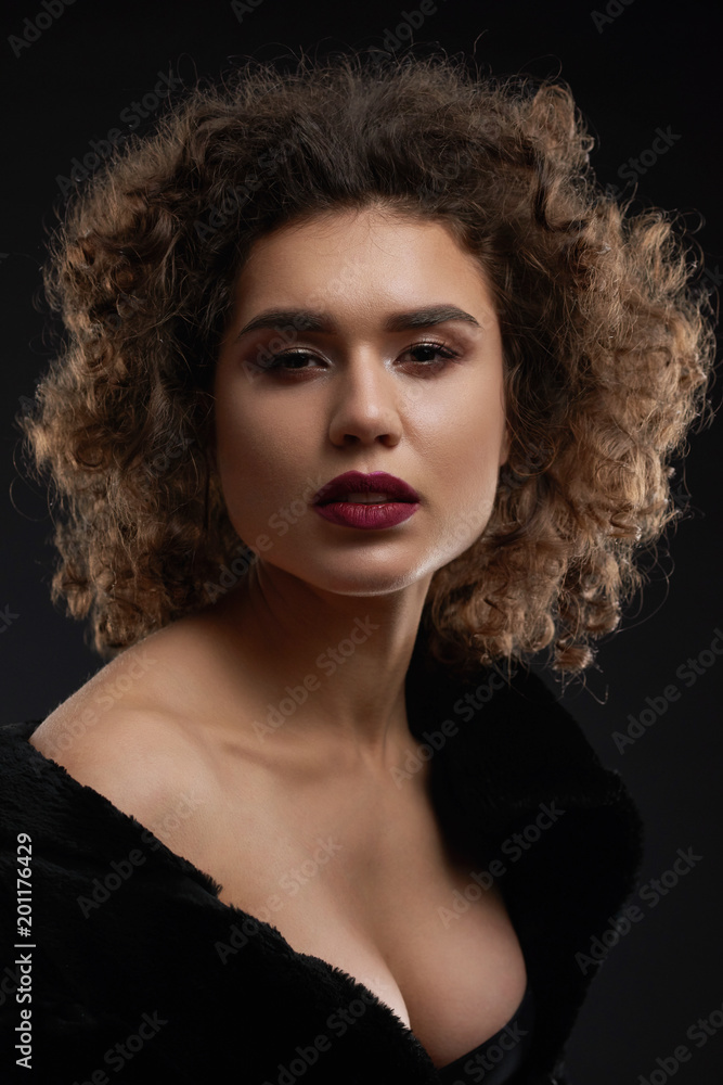 Sexy curly woman posing in the studio and looking at camera. Looking beatiful and passionate. Wearing light day make up and saturated violet lipstick on plump lips. Black background.
