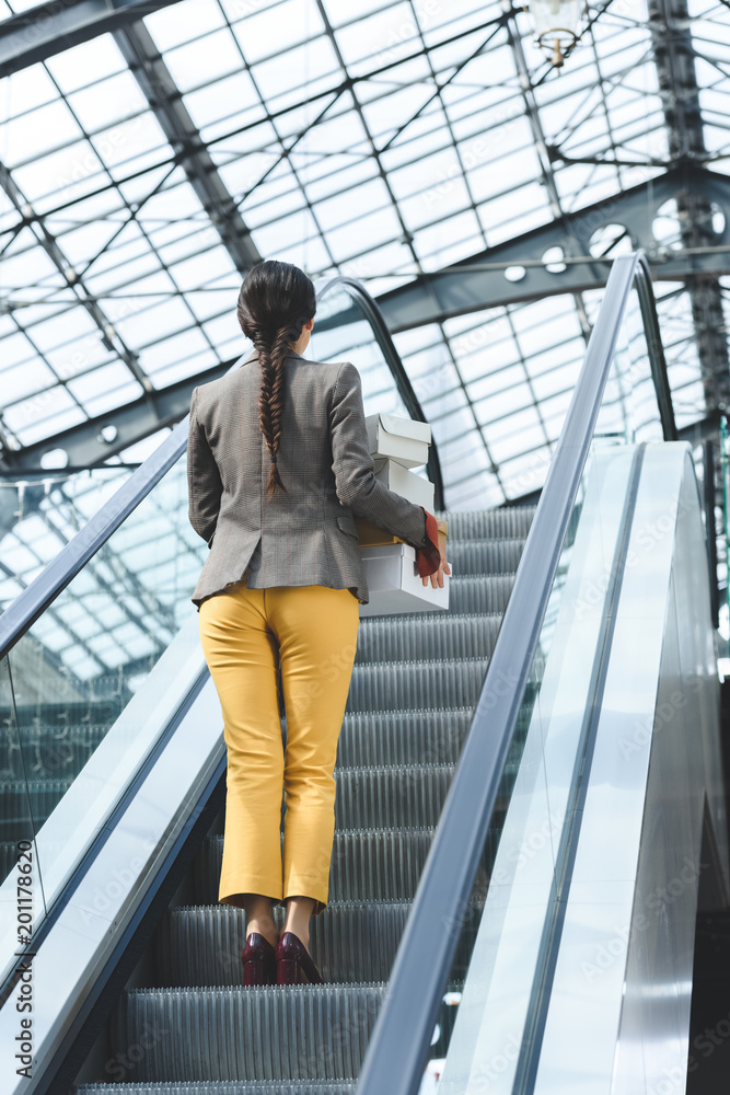 back view of woman standing with shopping boxes on escalator in mall