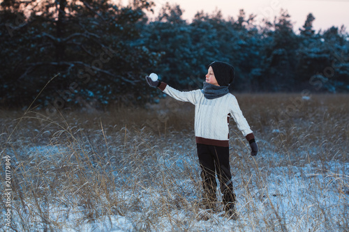 a boy in a sweater and a balloon plays snowballs on a snow-covered glade among the fir trees at sunset