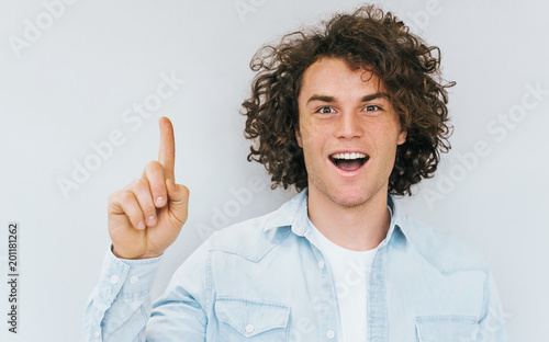 Horizontal portrait of curly hair amazed handsome man has excited expression, index finger up, recieve good idea for new project work. Happy pleased male isolated on white studio wall. Copy space