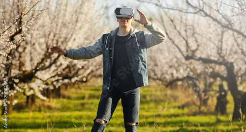 Young the guy of virtual reality glasses walk goes on the blossoming garden. Outdoor.