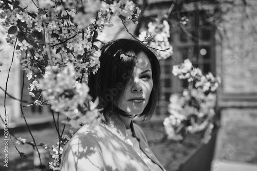 Beautiful pensive brunette girl with short hair is in a flowery spring garden with a cherry tree. The trees are covered with white delicate flowers. Black and white photo.