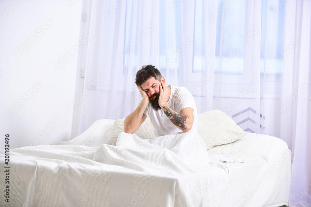 Man sitting on bed, falling asleep while lean on hand, white curtains on background. Handsome tired man leaning on his hand and sitting with closed eyes on his bed while getting up in the morning. Man sitting on bed, falling asleep while lean on hand, white curtains on background. Handsome tired man leaning on his hand and sitting with closed eyes on his bed while getting up in the morning.