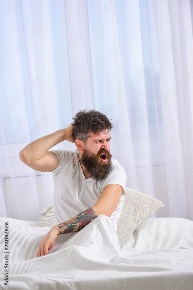 Hangover concept. Macho with beard and mustache suffers from hangover. Man in shirt sits on bed awake, white curtain on background. Guy on yawning face waking up in morning. Hangover concept. Macho with beard and mustache suffers from hangover. Man in shirt sits on bed awake, white curtain on background. Guy on yawning face waking up in morning.
