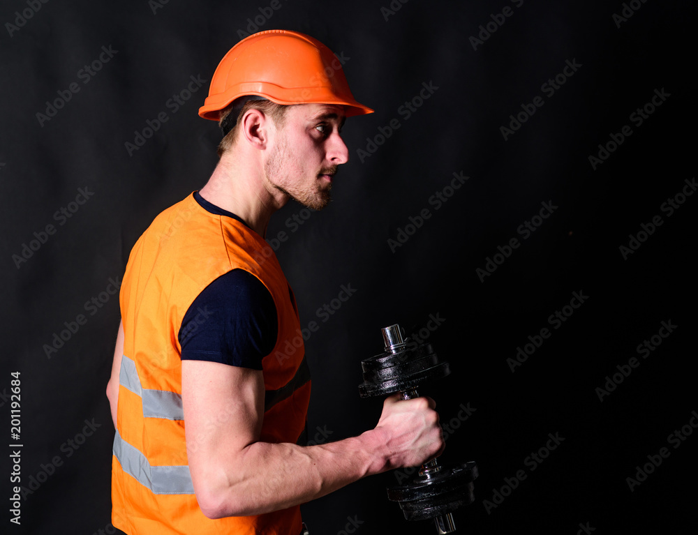 Repairman holds dumbbell in his right hand, black background. Strong builder concept. Builder in helmet working out, copy space. Worker, contractor, builder on strict face with muscular biceps. Repairman holds dumbbell in his right hand, black background. Strong builder concept. Builder in helmet working out, copy space. Worker, contractor, builder on strict face with muscular biceps.