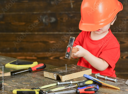 Kid boy hammering nail into wooden board. Toddler on busy face plays at home in workshop. Handcrafting concept. Child in too big helmet cute playing as builder or repairer, repairing or handcrafting.