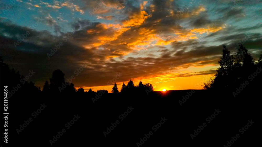 Dramatic red sunset / sunrise with cirrus clouds over rural field with silhouettes of forest trees and horizon line.