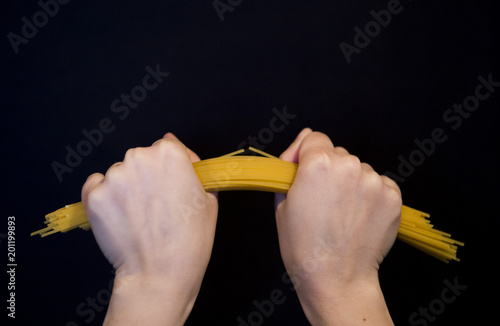  female hands clenching in fists bend, trying to break in half a bunch of raw long yellow thin spaghetti pasta on a black background