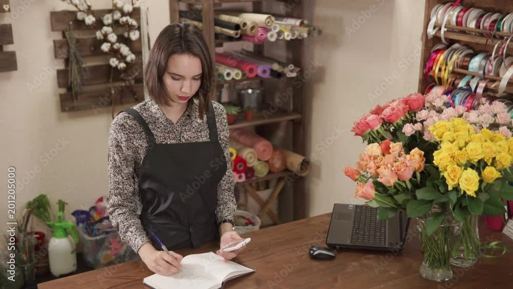 Female shop assistant is calculating using mobile phone, in a trading hall of flower shop. She is making notes in a log, notebook and vases with rose in a table Female shop assistant is calculating using mobile phone, in a trading hall of flower shop. She is making notes in a log, notebook and vases with rose in a table