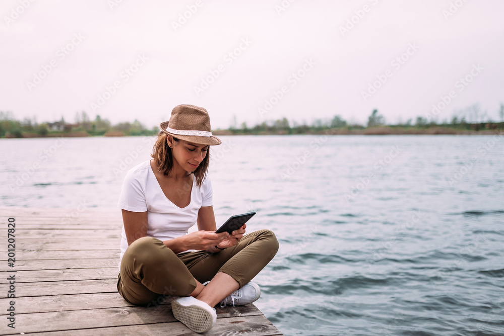 Woman with hat sitting near water and using tablet.