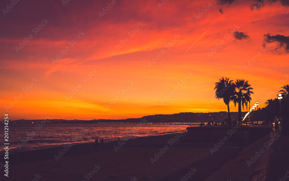 Beautiful pink, red and yellow colored sunset sky with silhouettes of people and palm trees at seaside in Nice, Cote d'Azur, France