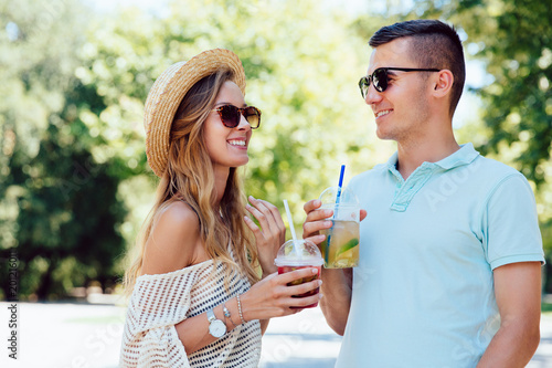 Cheerful beautiful couple having fun together, drinking fresh beverage outdoors, talking to each other. Dressed in summer clothes, in sunglasses.