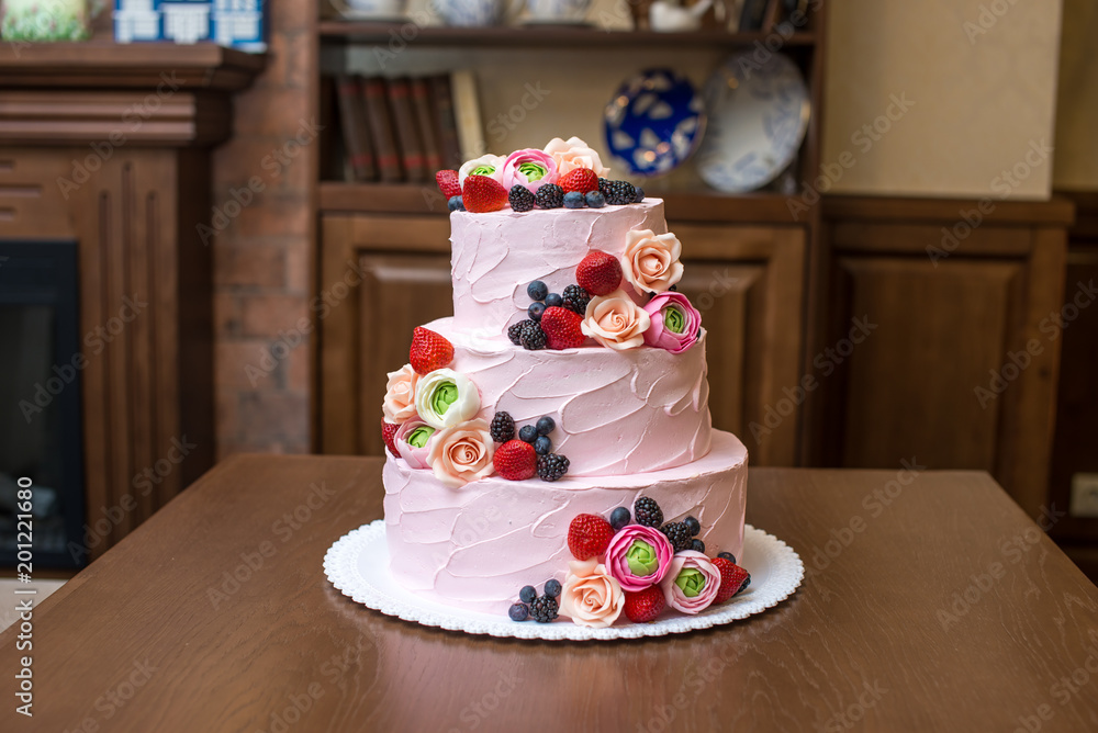 Fototapeta premium Three tiered pink wedding cake decorated with berries and flowers. Concept patisserie floristic from sugar mastic