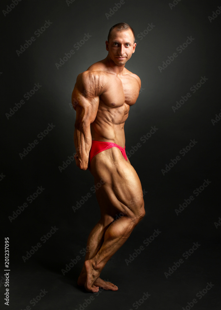 Studio shot of male bodybuilder posing, showing front muscles, huge triceps and legs, with black background.