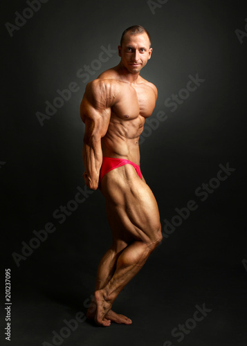 Studio shot of male bodybuilder posing, showing front muscles, huge triceps and legs, with black background.