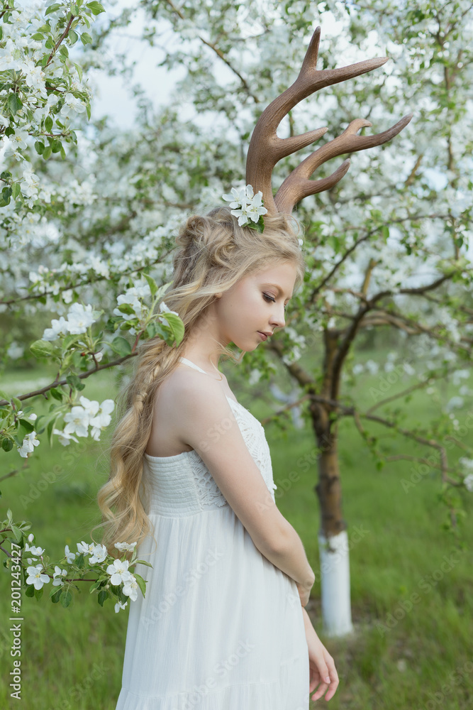 Naklejka premium Teen beautiful blonde girl wearing white dress with deer horns on her head and white flowers in hair stays in a spring blooming garden of trees covered with white flowers.
