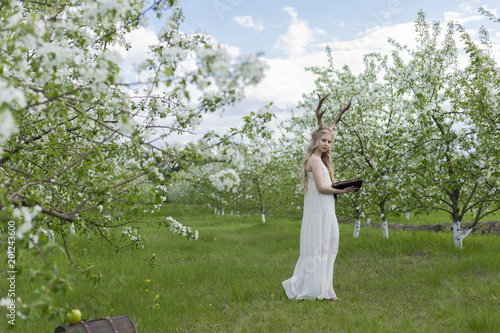 Teen beautiful blonde girl wearing white dress with deer horns on her head and white flowers in hair stays in a spring blooming garden holds an open antique book in her hands © Алексей Торбеев