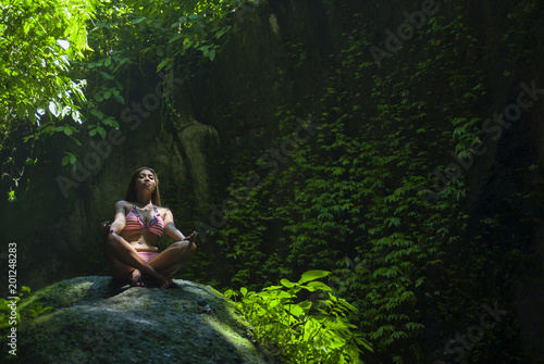 young beautiful Asian woman practicing Yoga posing sitting in lotus position meditating over a stone in a stunning natural landscape full of vegetation © TheVisualsYouNeed