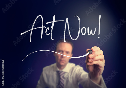 A businessman writing a Act Now concept with a white pen on a clear screen.