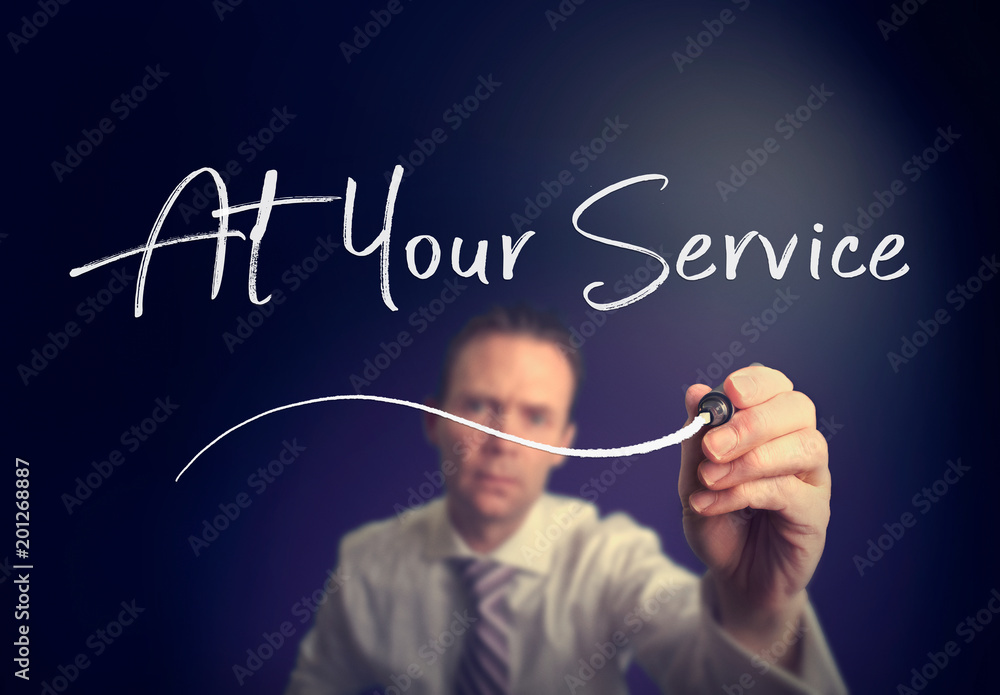 A businessman writing a At Your Service concept with a white pen on a clear screen. A businessman writing a At Your Service concept with a white pen on a clear screen.