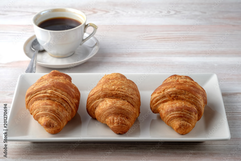 Three croissants on a rectangular plate and a cup of black coffee on a light table texture. Place for your text. Three croissants on a rectangular plate and a cup of black coffee on a light table texture. Place for your text.