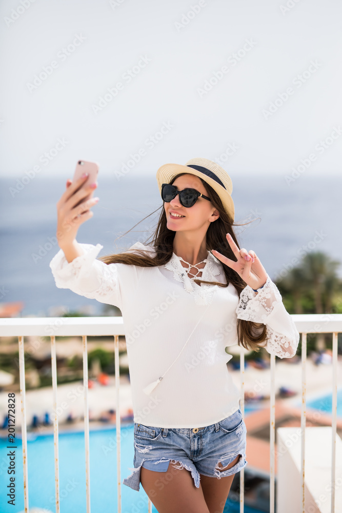 Cute young woman making selfie on ocean beach, wearing boho outfit and funny sunglasses, straw hat, sending peace on sea background.