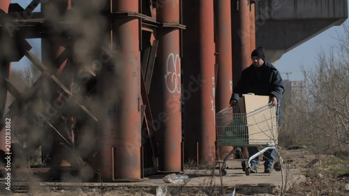 Poor homeless bearded man in jacket and hat walking in cold autumn weather uner the bridge, pushing shopping cart with belongings. Beggar male gathering plastic bottle from the ground. photo