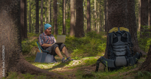 digital nomad, young caucasian man working on his laptop sitting on a camping chair in the woods, backpacking,