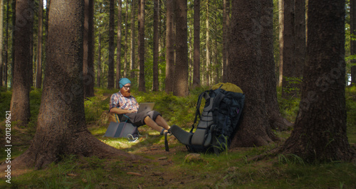 digital nomad, young caucasian man working on his laptop sitting on a camping chair in the woods, backpacking,