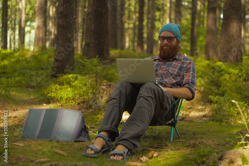 digital nomad, young caucasian man working on his laptop sitting on a camping chair in the woods, backpacking,