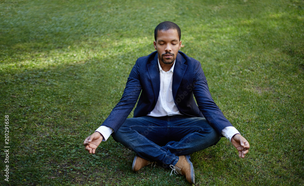 Young american african businessman in informal clothes meditating in lotus pose taking a deep breath at green grass.Business yoga and stress free environment.Peace of mind concept.Blurred Young american african businessman in informal clothes meditating in lotus pose taking a deep breath at green grass.Business yoga and stress free environment.Peace of mind concept.Blurred