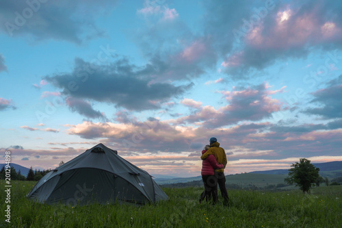 adventure hiking couple, man and woman standing next to a tent pitched on a meadow looking at clouds during sunset