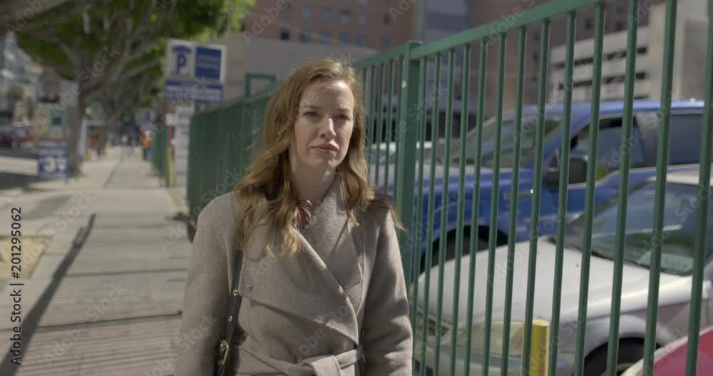 Medium shot, smartly dressed business woman in coat walking past fenced parking lot in Downtown Los Angeles. Warm morning light. Hand-held, moving camera, slow motion 4K recorded at 60fps Medium shot, smartly dressed business woman in coat walking past fenced parking lot in Downtown Los Angeles. Warm morning light. Hand-held, moving camera, slow motion 4K recorded at 60fps